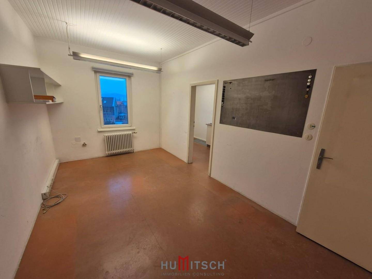 Kleinerer Büroraum mit einem Fenster, Heizkörper, einer Tür und einer dunklen Tafel an der Wand.