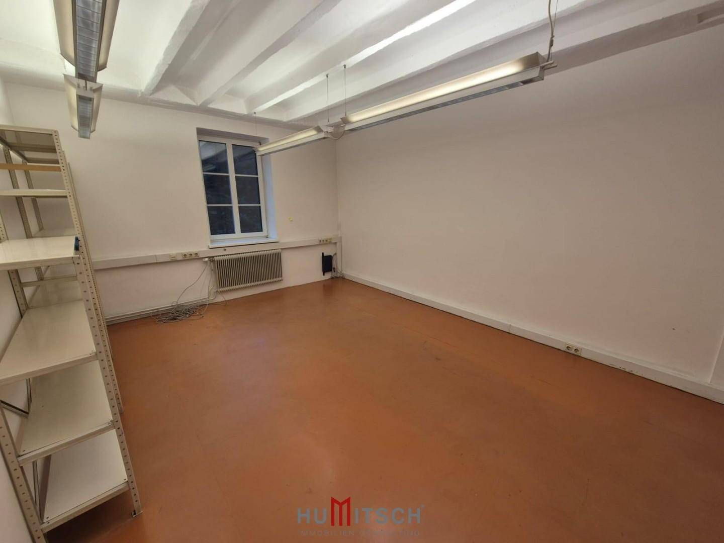 Leerer Raum mit einem Fenster, Heizkörper, Metallregal und sichtbaren Deckenbalken.