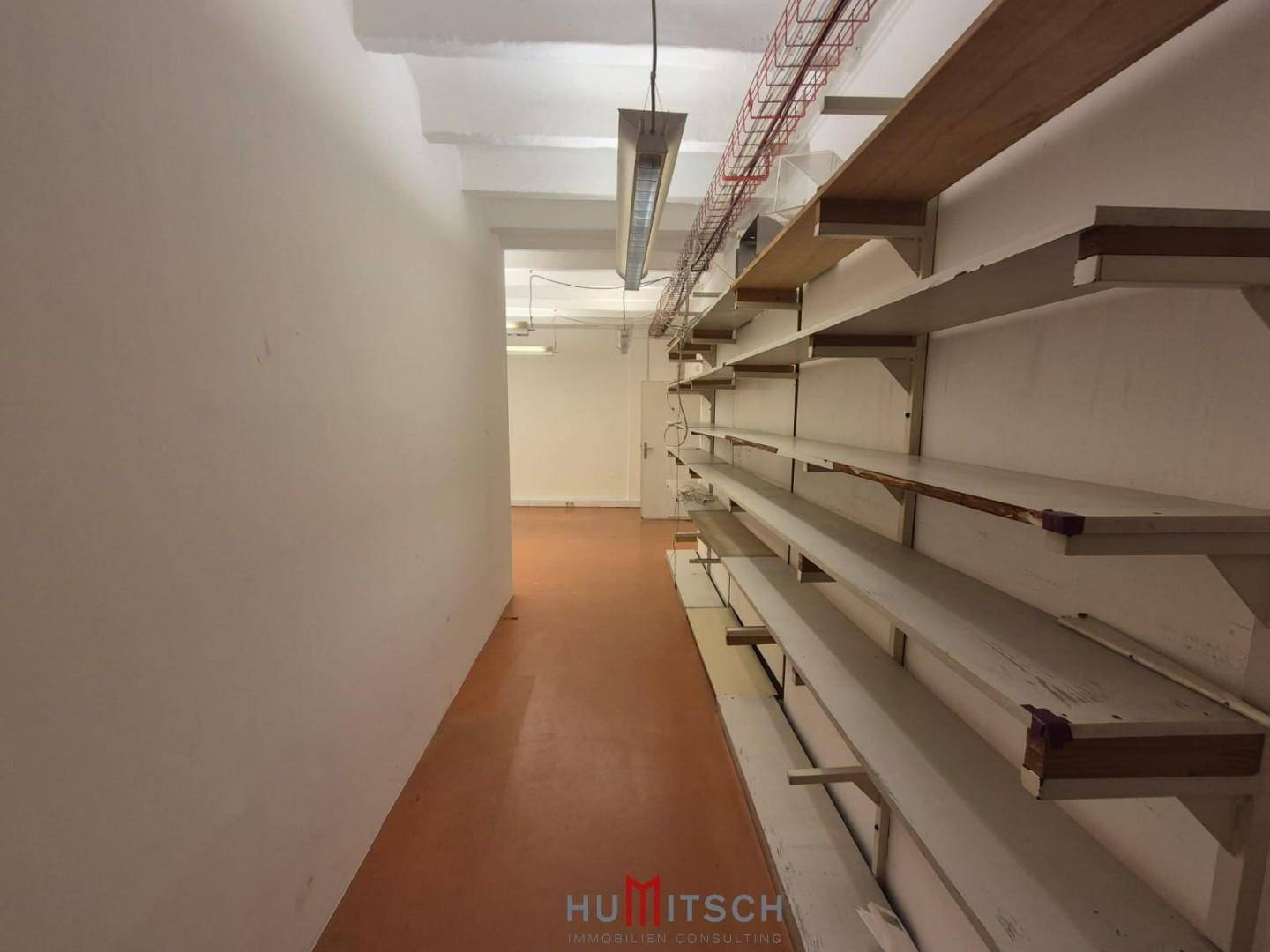 Langer Lagerraum oder Flur mit Regalen an der Wand und einfacher Deckenbeleuchtung.