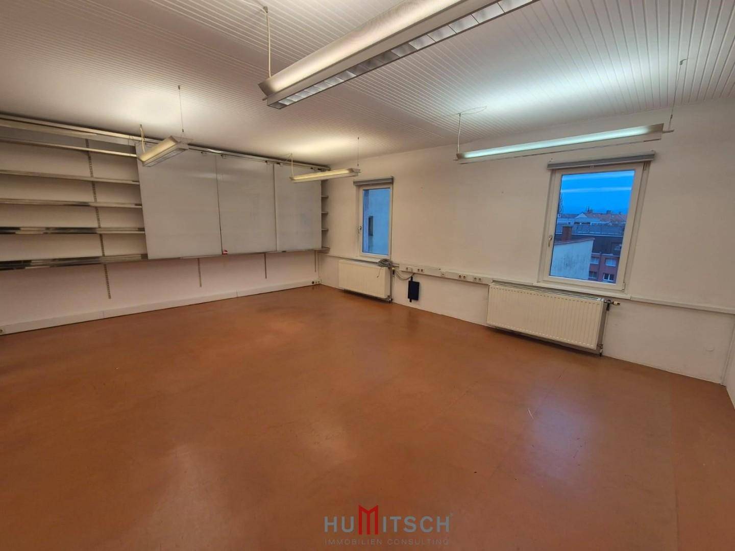Heller Büroraum mit Regalen, Whiteboard, zwei Fenstern und funktionaler Deckenbeleuchtung.