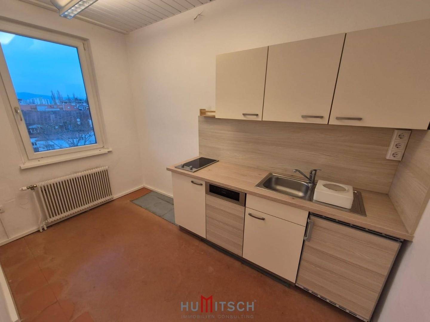 Kleine Küchenzeile mit hellen Holzschränken, Spüle, Kochfeld und Geschirrspüler, neben einem Fenster.