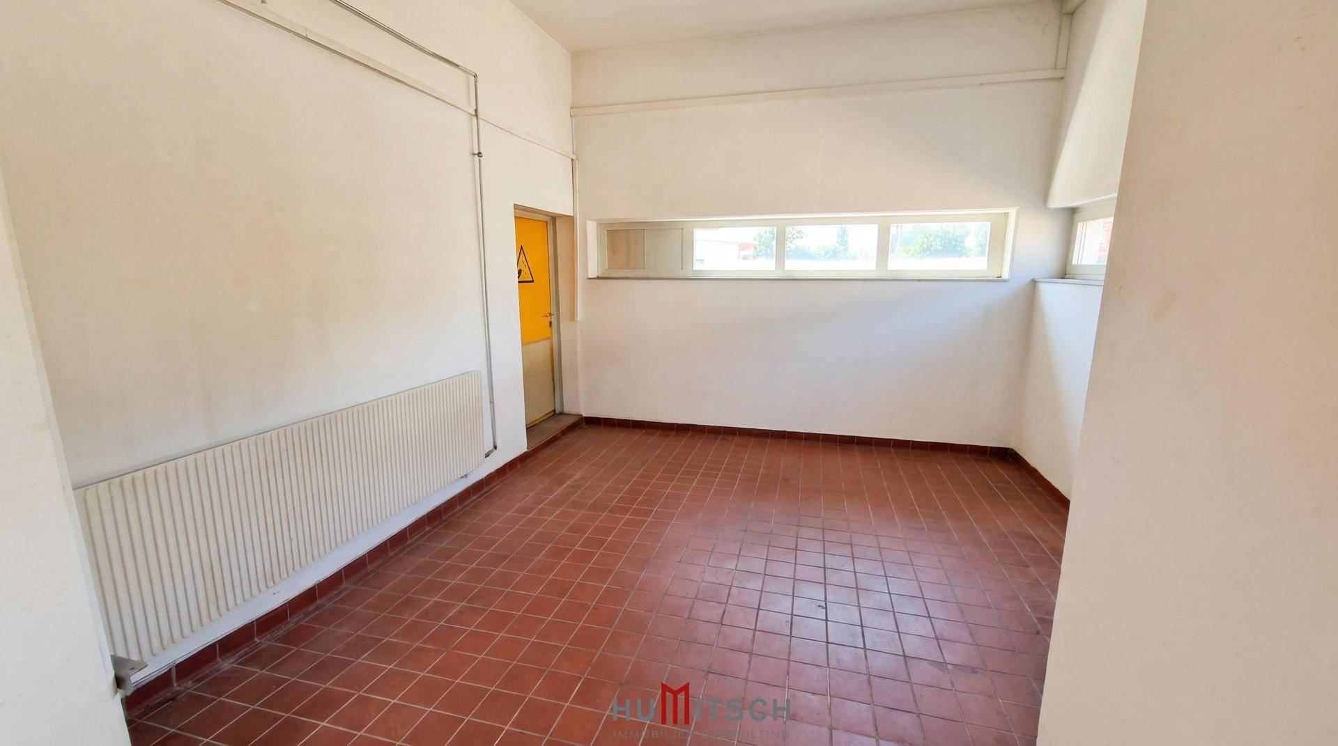 Kleiner Büroraum mit Terrakottafliesen, Heizkörper und Fenster, ideal für administrative Aufgaben.