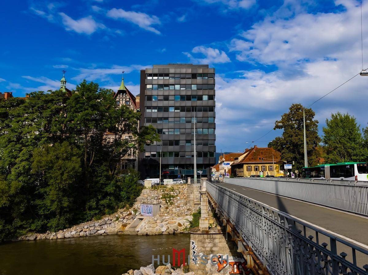 Ansicht des modernen Gebäudes von der Brücke aus, mit Fluss und historischer Architektur im Hintergrund.