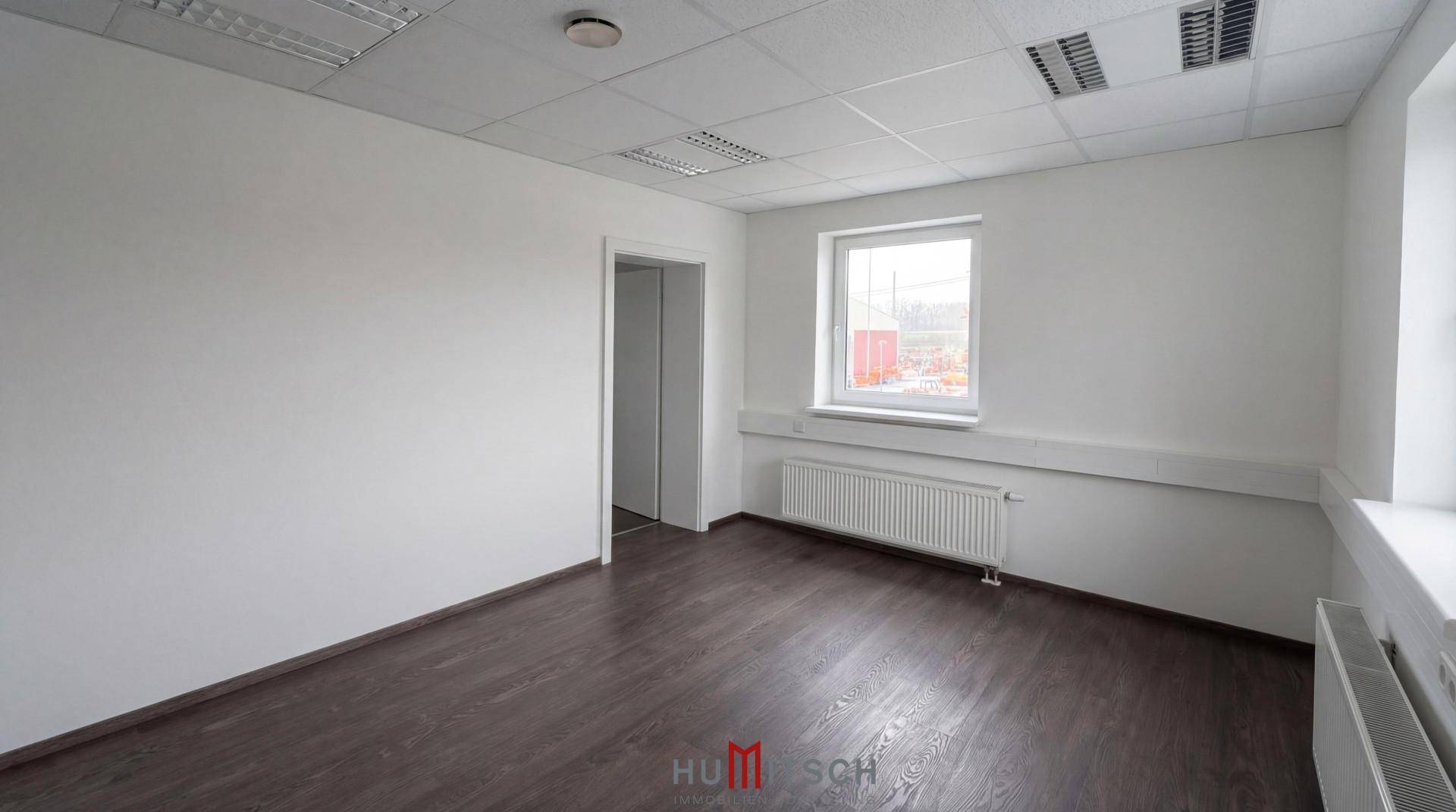 Leerer Büroraum mit dunklem Boden, weißen Wänden, einem Fenster und Zugang zu weiteren Räumen.