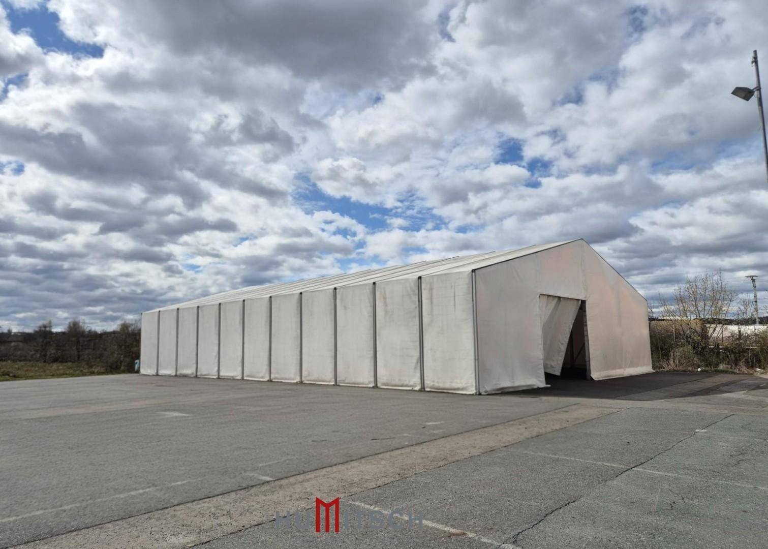 Großes weißes Zelt oder temporäre Lagerhalle auf einem asphaltierten Gelände unter bewölktem Himmel.