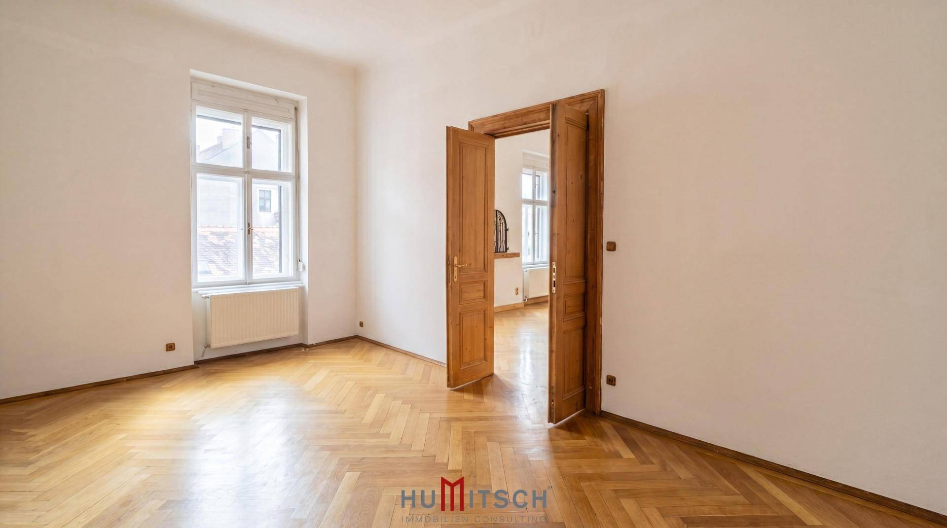 Geräumiges Zimmer mit Fischgrätparkett, weißen Wänden und einer hohen Holztür.