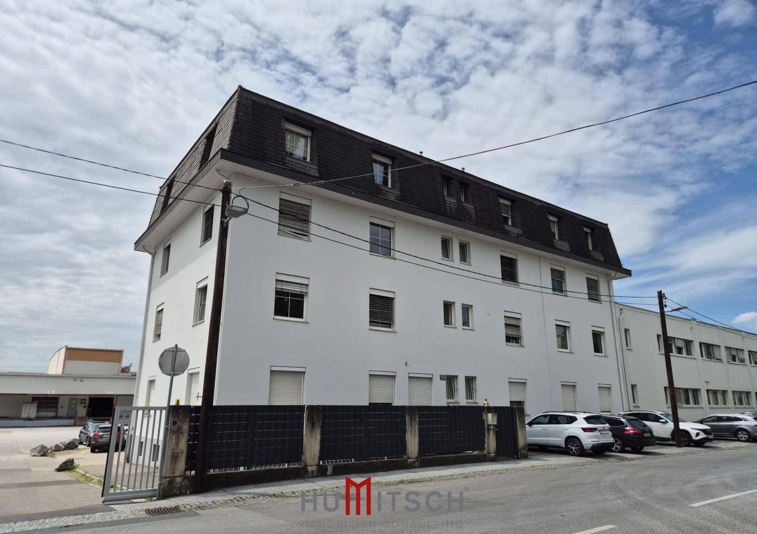 Ansicht des Gebäudes von der Straßenseite mit Parkplätzen und einem Zaun vor der weißen Fassade.
