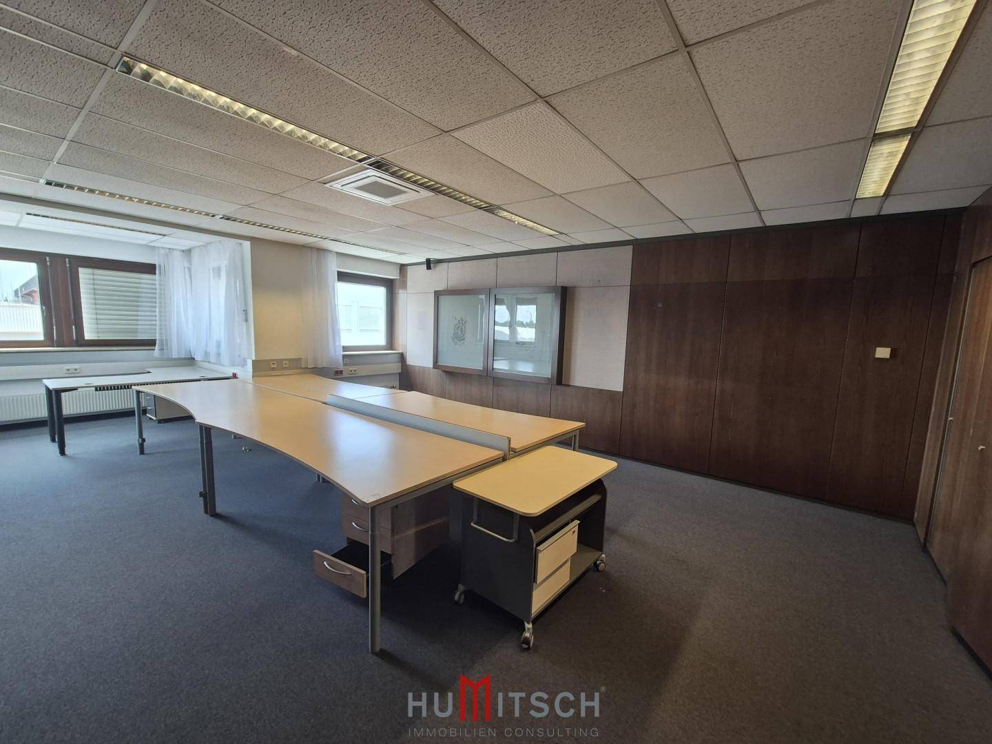 Büro mit Teppichboden, Holzvertäfelung an einer Wand und mehreren Schreibtischen mit Bürostühlen.