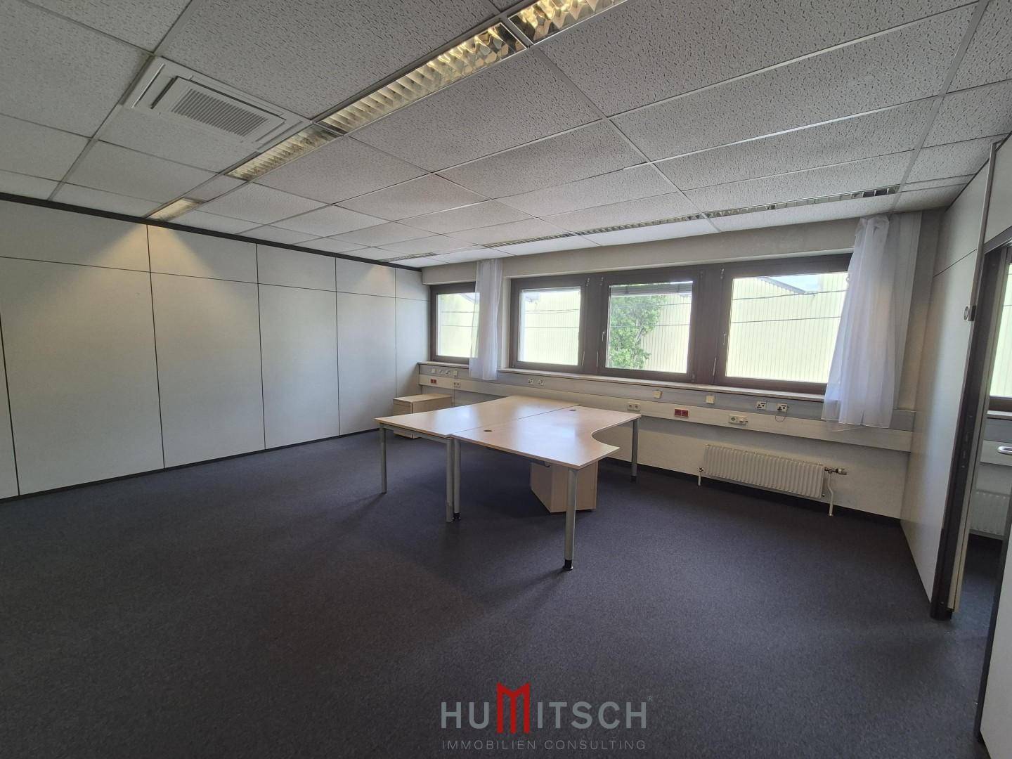 Helles Büro mit Teppichboden, weißen Wänden und einem Fensterbereich mit einem einzelnen Schreibtisch.