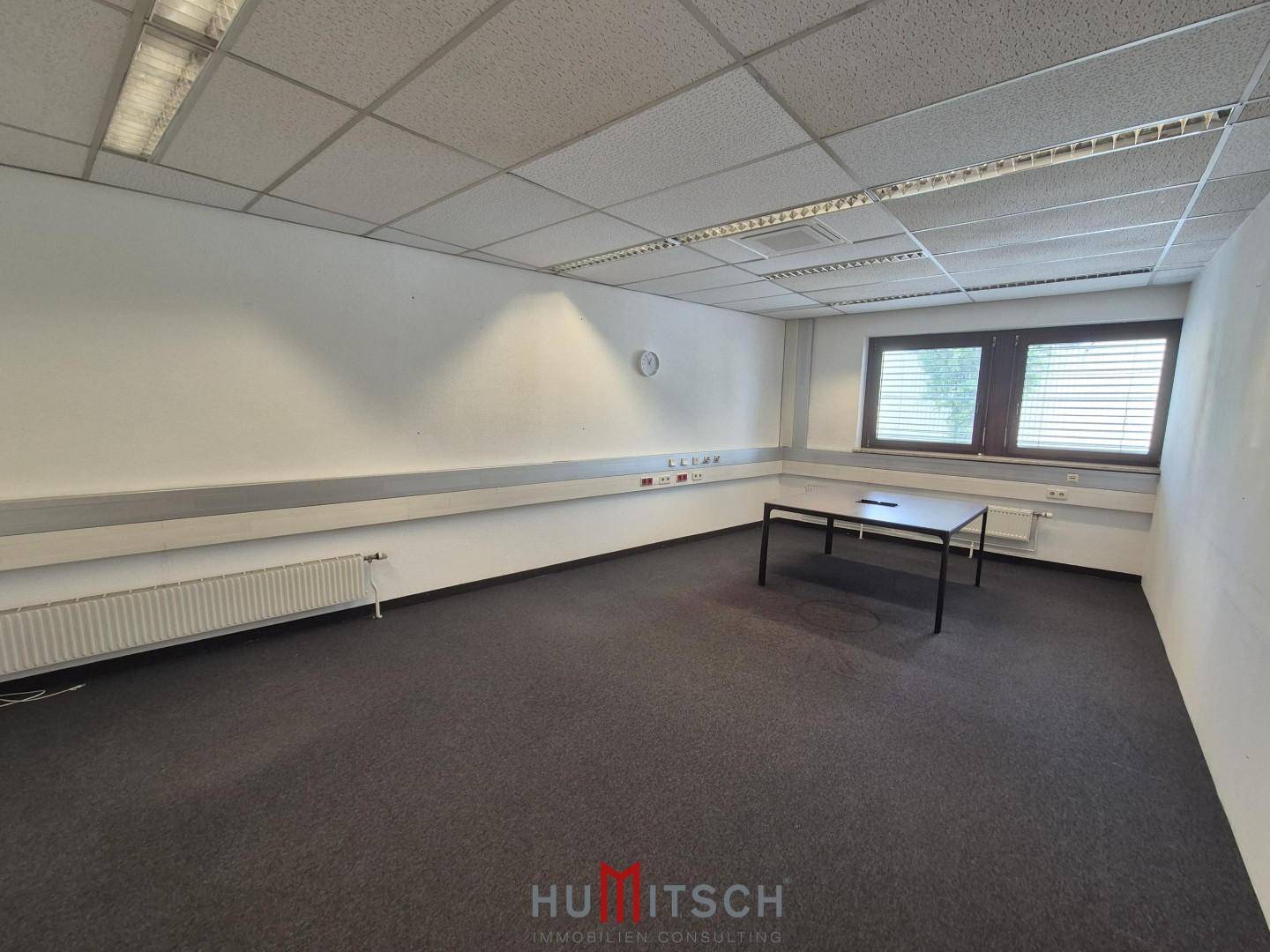 Einfaches Büro mit Teppichboden, weißen Wänden und einem kleinen Tisch unter einem Fenster.