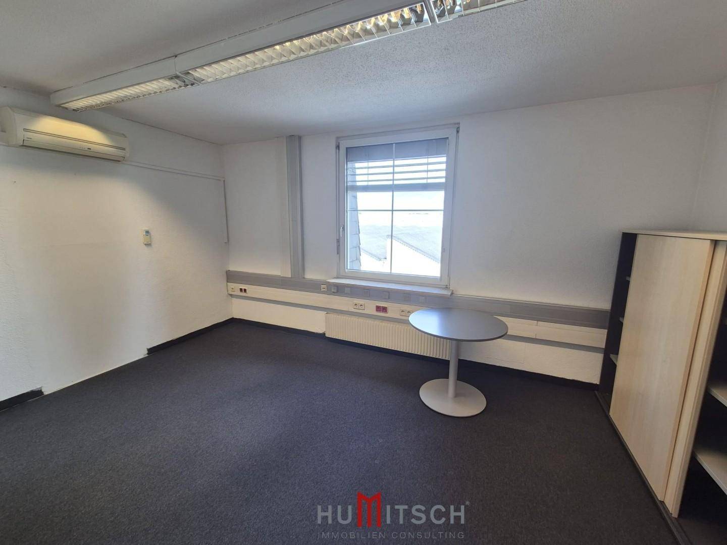Büro mit Teppichboden, einem runden Tisch und einem Klimagerät an der Wand.