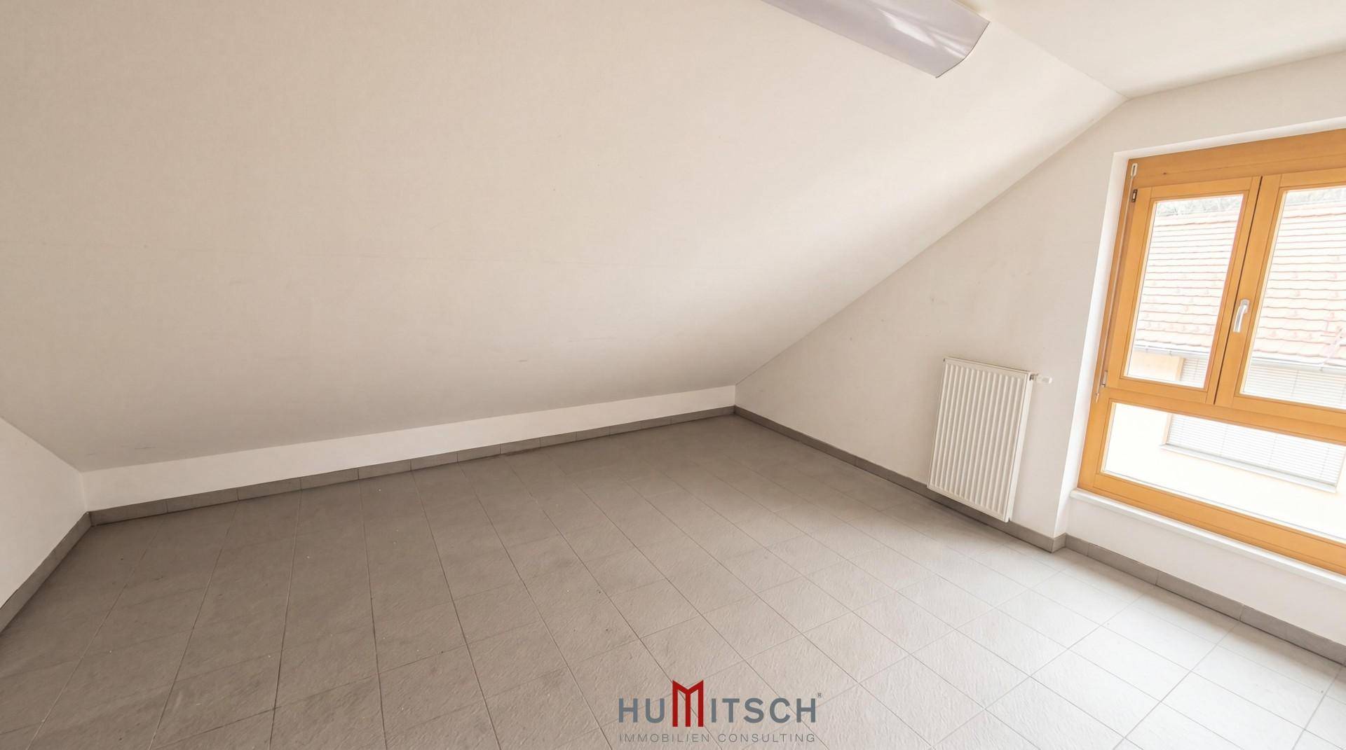 Dachbodenraum mit schrägen Wänden, Fliesenboden und einem Fenster, das den Raum mit Tageslicht versorgt.