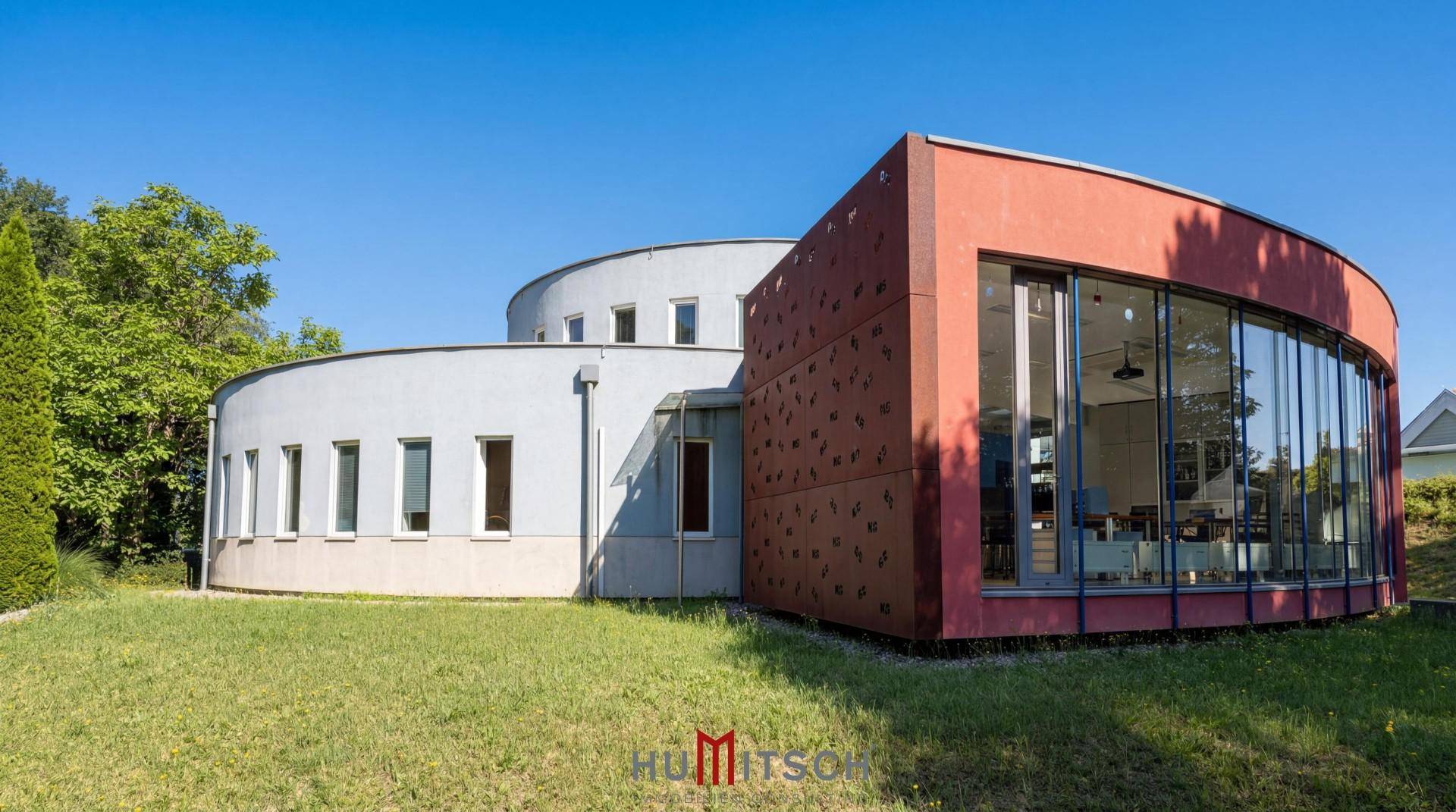 Moderne Architektur mit geschwungener Fassade und großen Glasfronten, umgeben von Grünflächen.
