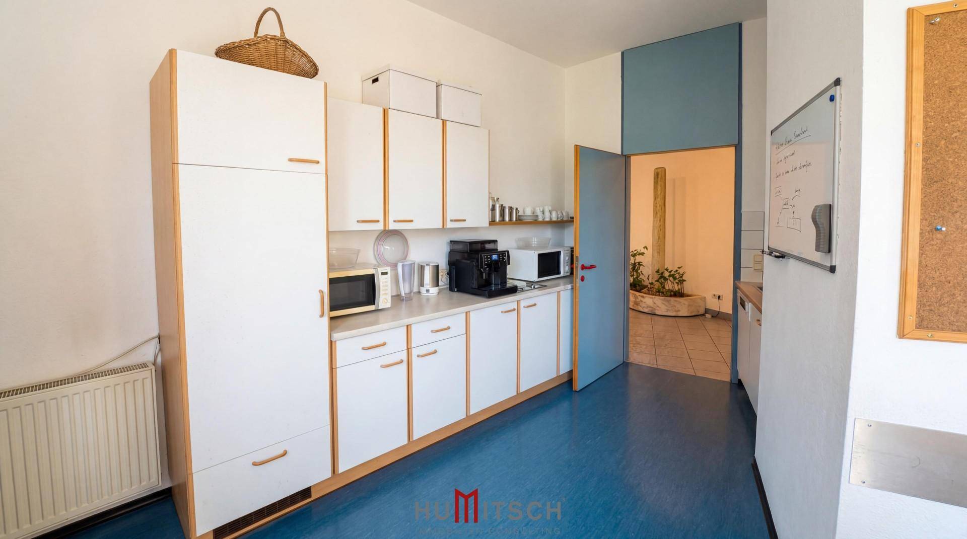 Funktionale Küche mit weißen Schränken, Mikrowelle, Kaffeemaschine und blauem Boden.