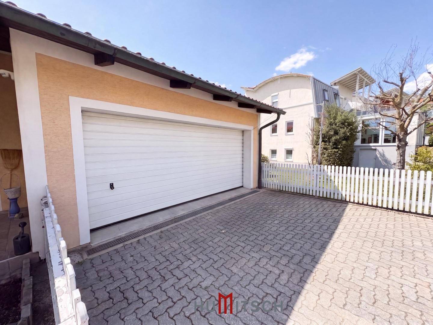Geräumige Garage mit weißem Tor und gepflasterter Zufahrt, umzäunt von einem Lattenzaun.