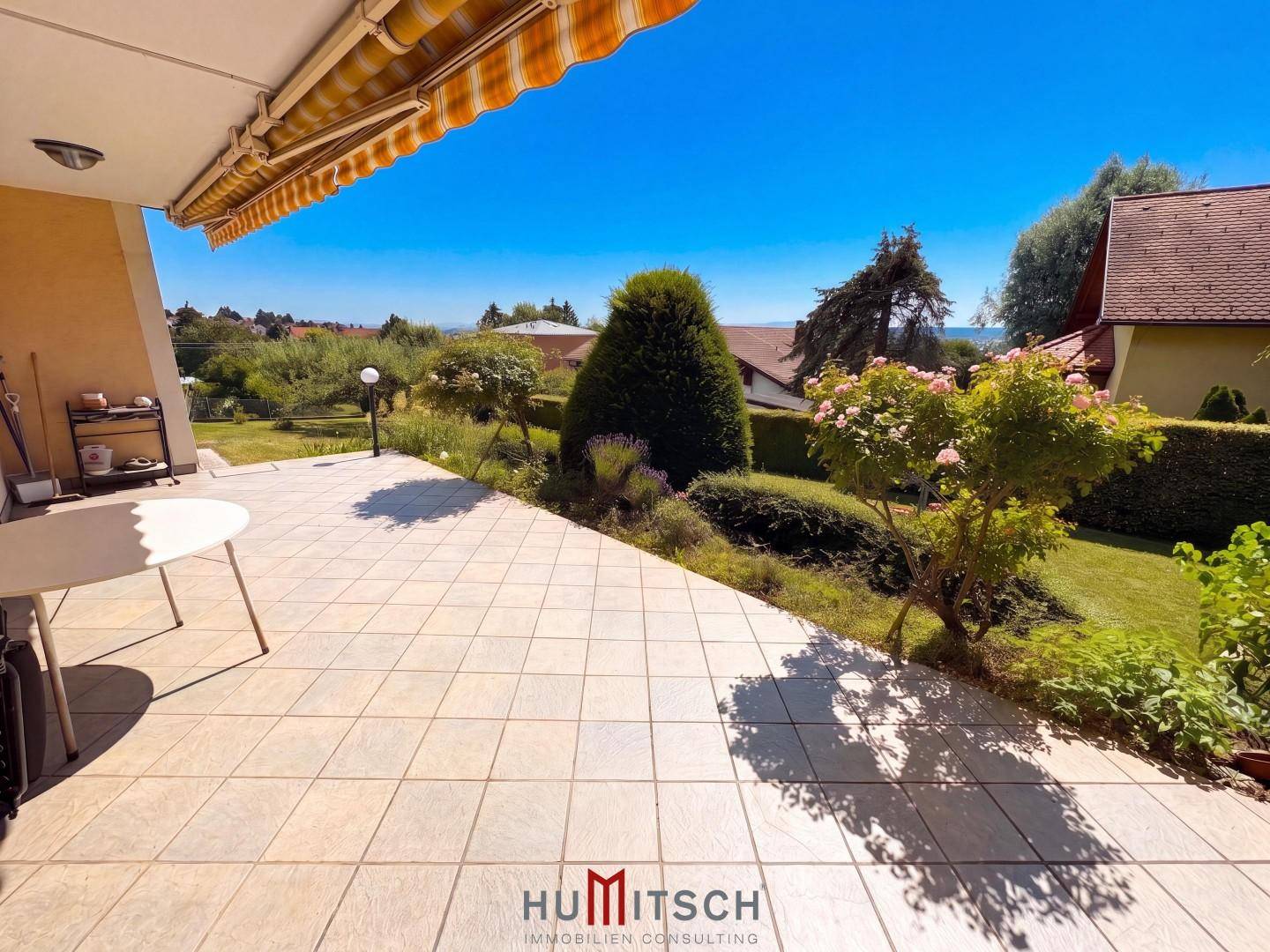 Große geflieste Terrasse mit Markise, Gartenmöbeln und Blick ins Grüne.