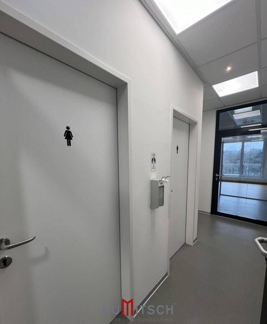 Heller Flur mit Zugang zu den Toiletten und einem Spender für Händedesinfektionsmittel.