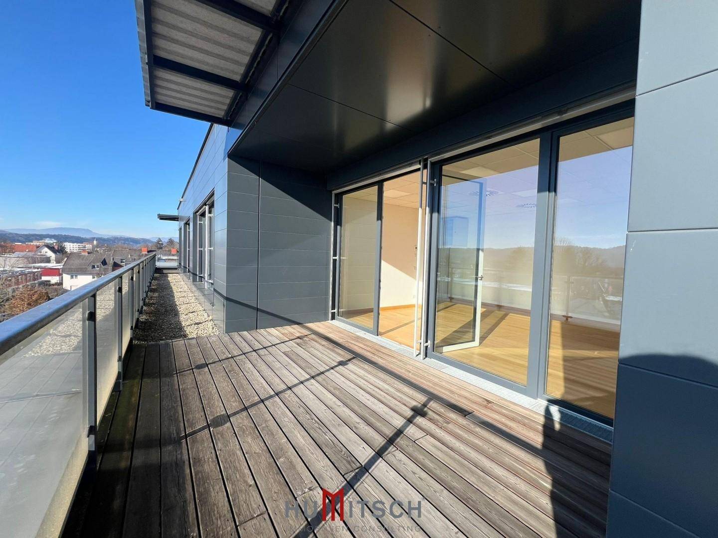 Geräumige Holzterrasse mit Metallgeländer und Blick auf die Umgebung unter blauem Himmel.