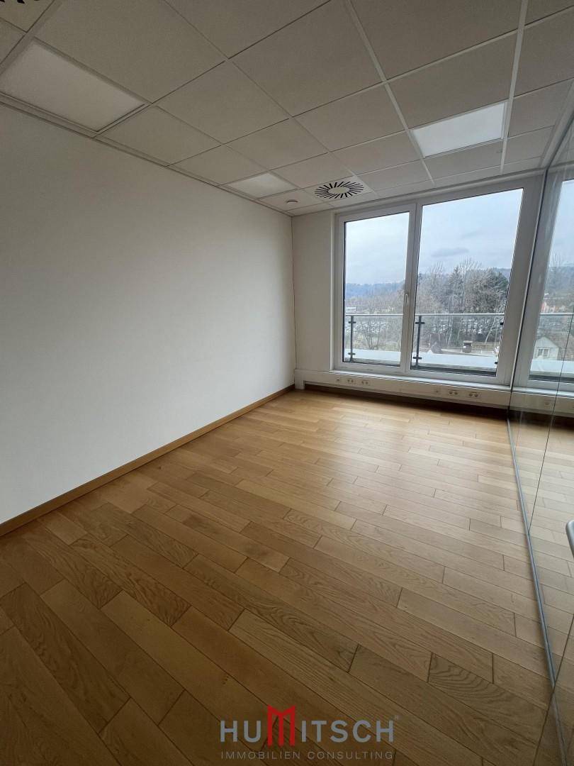 Leerer Büroraum mit Holzboden, abgehängter Decke und großen Fenstern, die den Raum erhellen.