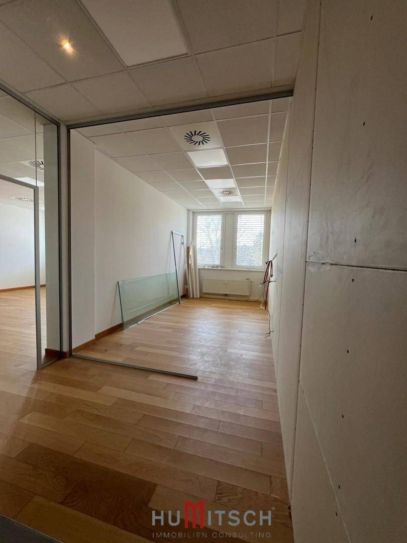 Büroflur mit Holzboden, abgehängter Decke und Fenstern am Ende, mit Baumaterialien.
