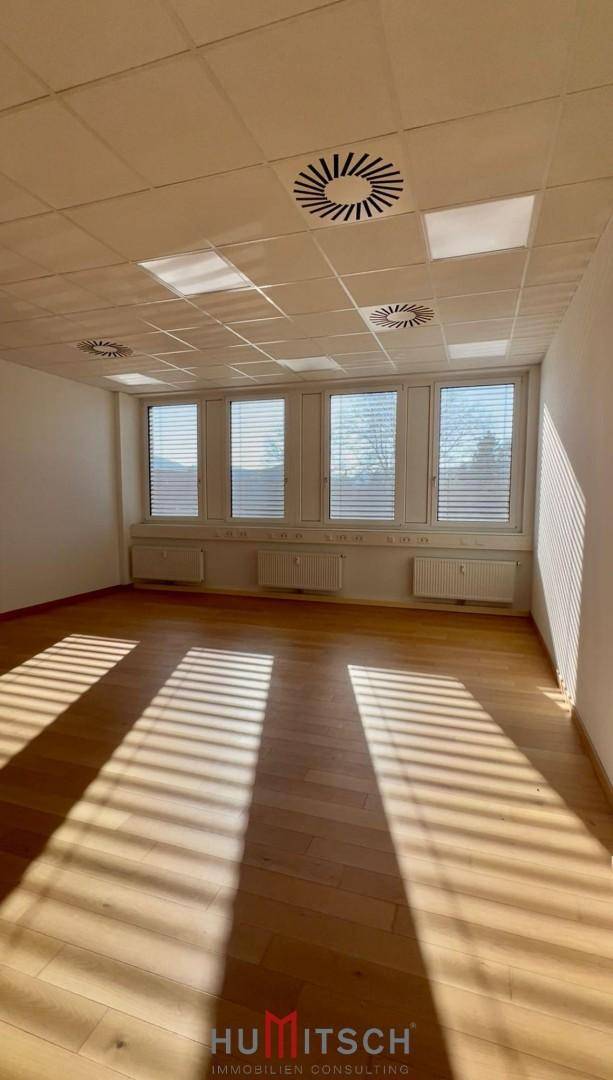 Heller Büroraum mit Holzboden, großen Fenstern mit Jalousien und abgehängter Decke.