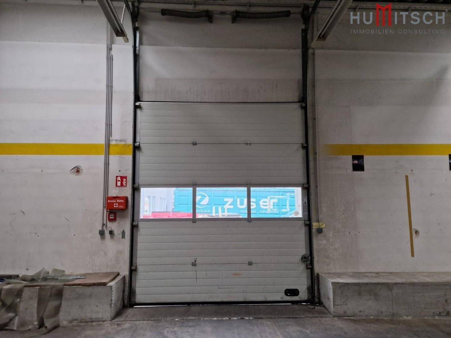 Innenansicht eines großen Rolltors mit Fenster, das zu einem Ladebereich führt, inklusive Feuerlöscher.