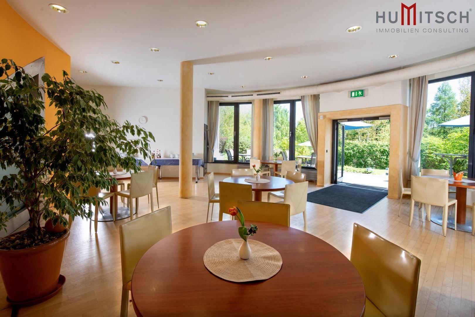 Heller Speisesaal mit Holzboden, runden Tischen und Zugang zu einer Außenterrasse mit Grünblick.