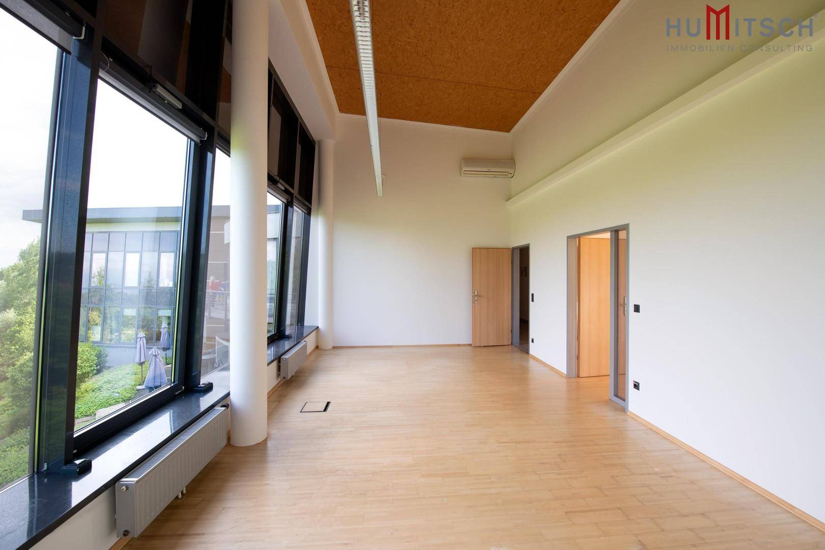 Großes Büro mit Holzboden, Klimaanlage und bodentiefen Fenstern, die viel Licht und Ausblick bieten.