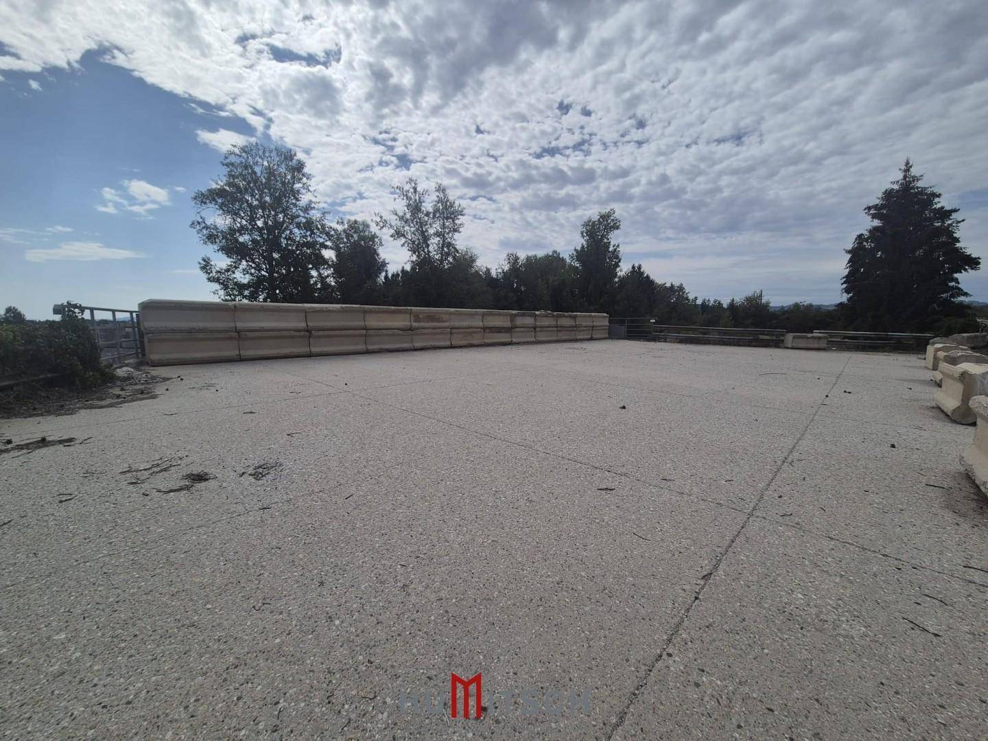 Große, ebene Betonfläche, umgeben von einer niedrigen Mauer und Bäumen unter bewölktem Himmel.