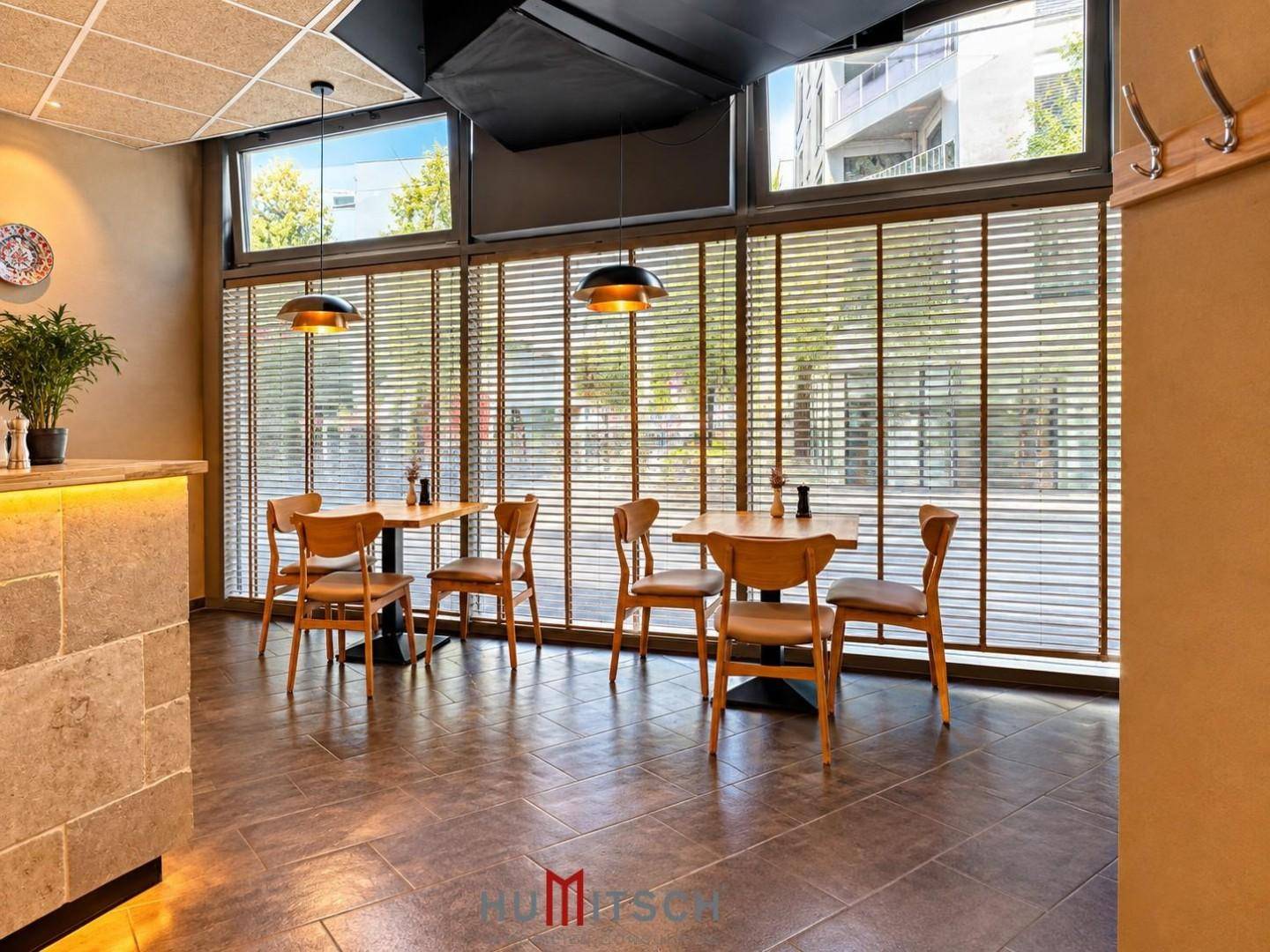 Gemütlicher Restaurantbereich mit Holztischen und Stühlen, großen Fenstern und Jalousien für Privatsphäre.