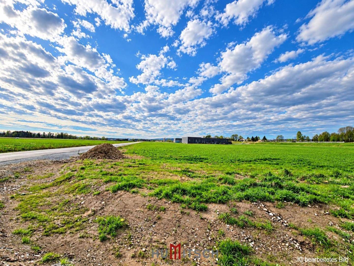 Weitläufige grüne Landschaft mit einem Erdhügel am Straßenrand und Gebäuden in der Ferne.