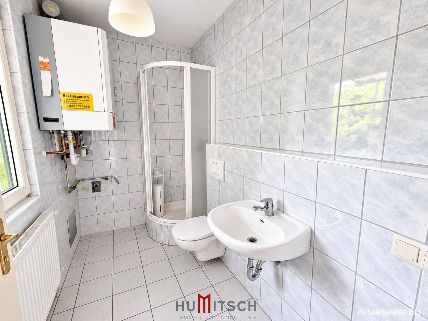 Badezimmer mit hellgrauen Fliesen, Duschkabine, WC und Waschbecken sowie einem Fenster.