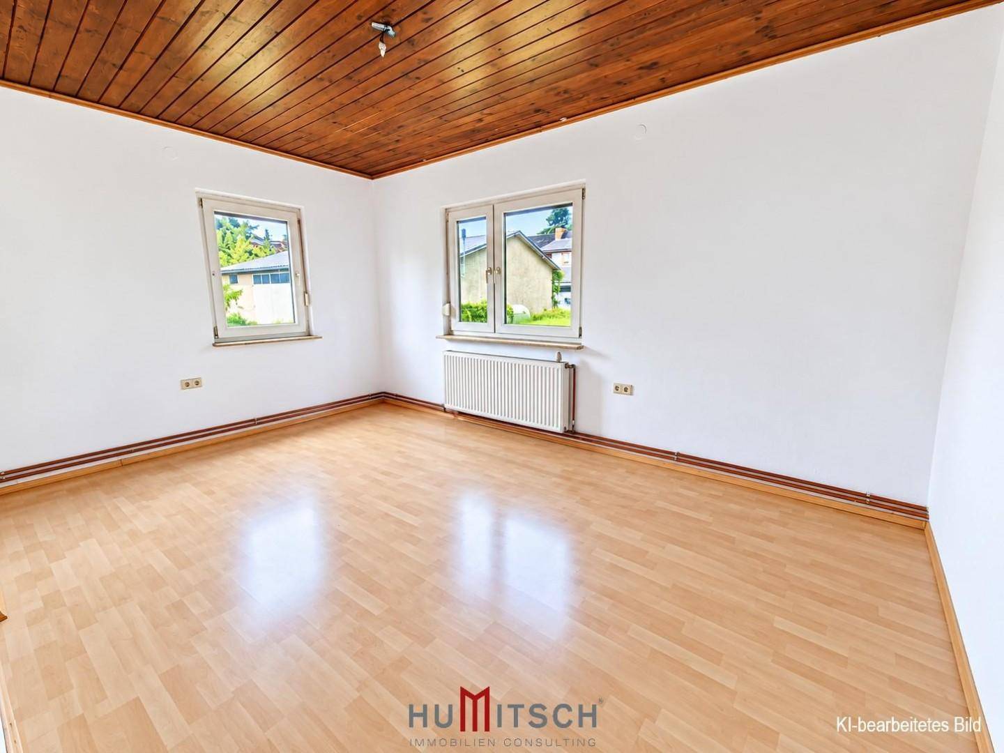 Leerer Raum mit Holzbalkendecke, Laminatboden und zwei Fenstern mit Blick ins Grüne.