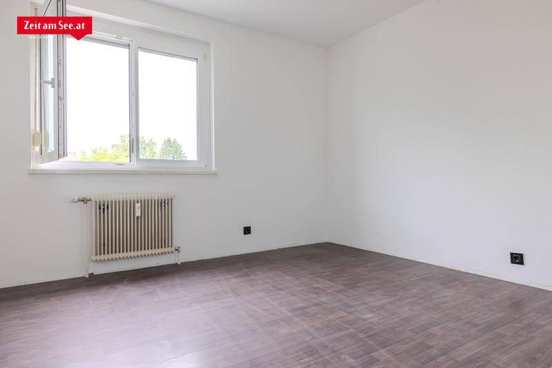 Leeres Zimmer mit hellem Laminatboden, weiß gestrichenen Wänden und großem Fenster.