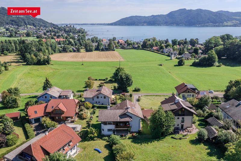 Luftaufnahme einer Wohnsiedlung mit grünen Feldern, See und Bergen im Hintergrund.
