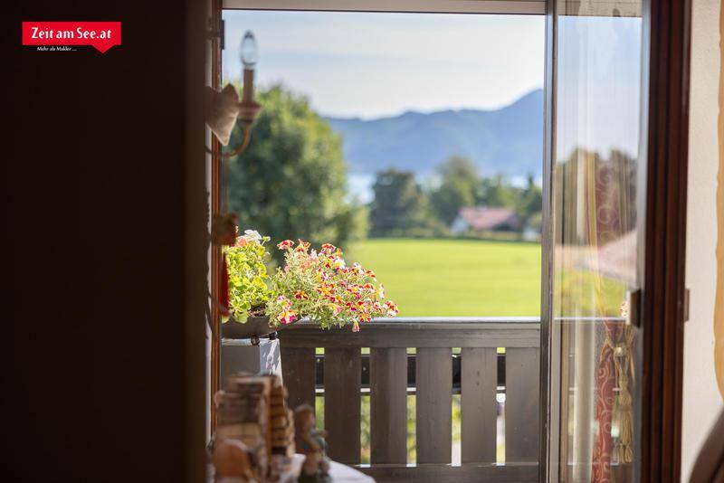 Blick von einem Innenraum auf einen Balkon mit Blumen und weitem See- und Bergblick.