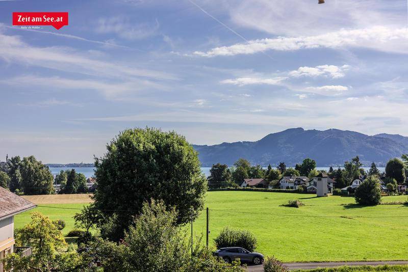Weitläufiger Blick über grüne Felder zum See und den umliegenden Bergen unter blauem Himmel.