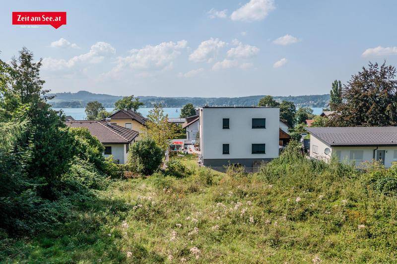 Blick über ein unbebautes Grundstück mit Häusern im Hintergrund und einem See in der Ferne.