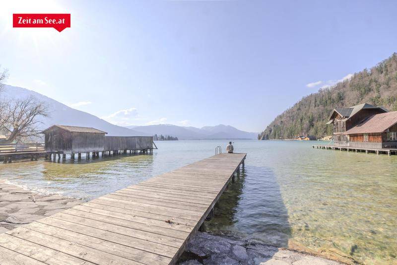 Holzsteg am Seeufer mit einer Person, die den weiten Blick über den See und die umliegenden Berge genießt.
