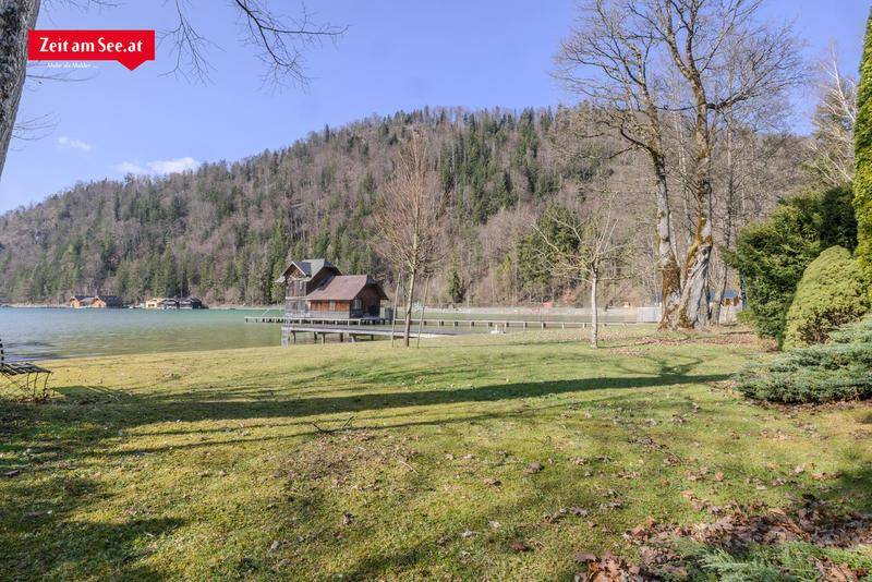 Weitläufige Rasenfläche am Seeufer mit Blick auf bewaldete Hügel, Bootshäuser und den klaren See.