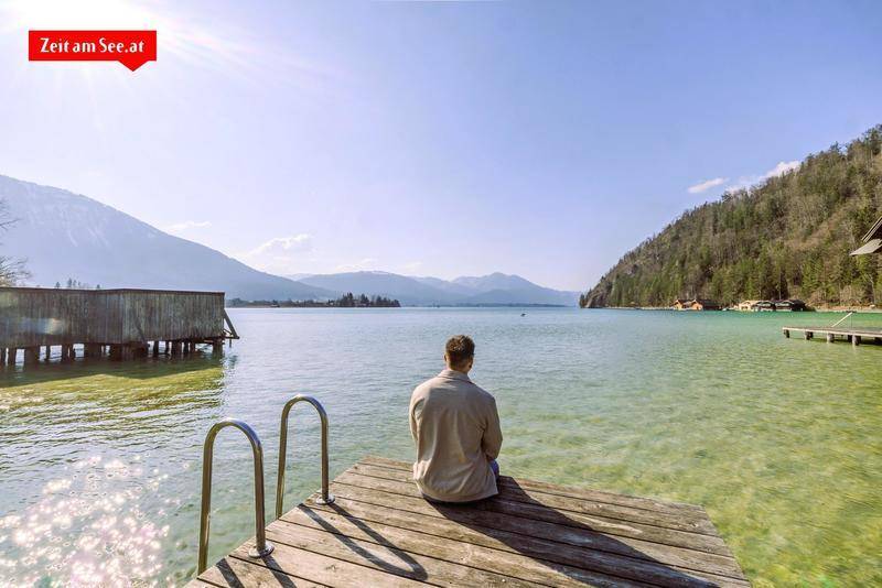 Idyllischer Blick auf einen klaren Bergsee mit Holzsteg und umliegenden Bergen unter blauem Himmel.