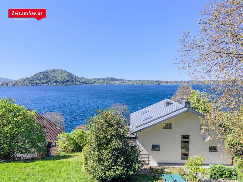 Modernes Haus mit Satteldach und großen Fenstern, eingebettet in eine grüne Landschaft mit Seeblick.