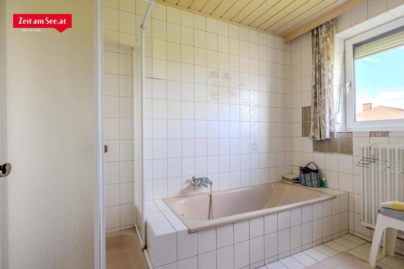 Badezimmer mit beiger Badewanne, weißen Fliesen und Holzdecke, ausgestattet mit einem Fenster.