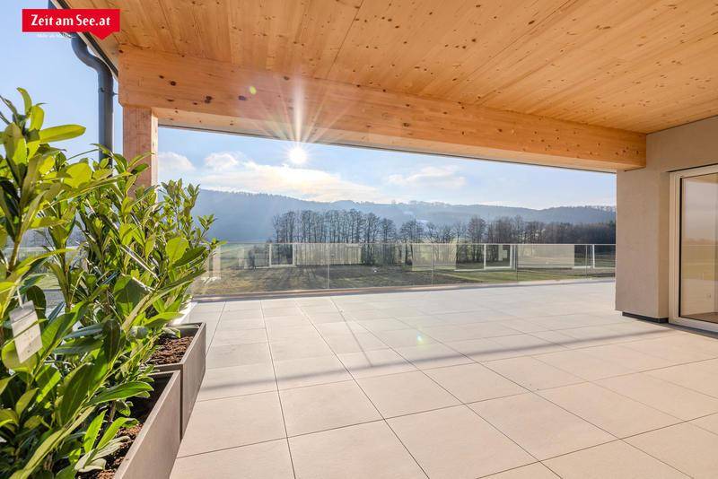 Sonnige Terrasse mit Glasgeländer und weitem Blick über grüne Hügel und einen See in der Ferne.
