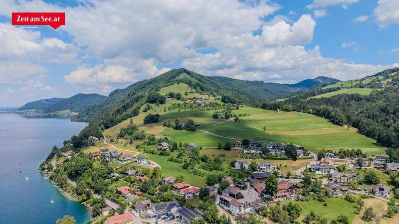 Panoramablick auf ein Dorf am Seeufer, umgeben von grünen Hügeln und Bergen unter blauem Himmel.
