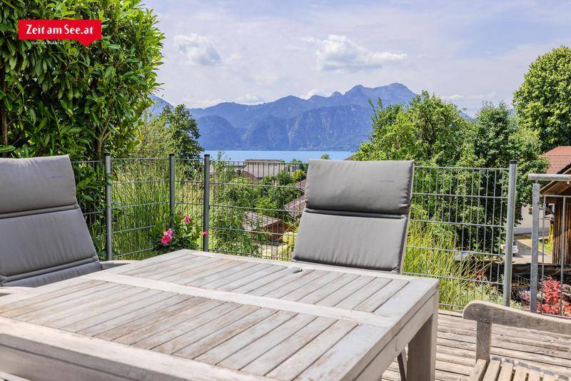 Terrasse mit Holztisch und Stühlen, umgeben von Grün und mit weitem Blick auf den See und die Berge.