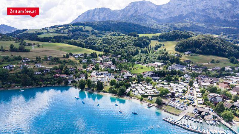Luftaufnahme einer malerischen Stadt am Seeufer mit Bergen im Hintergrund und zahlreichen Booten.