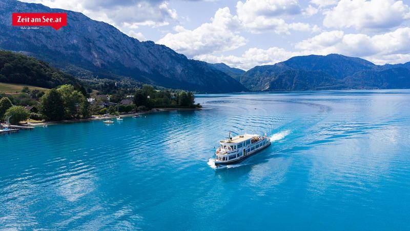 Malerischer See mit einem Passagierschiff, umgeben von majestätischen Bergen und üppiger Natur.