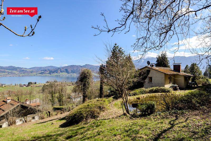 Haus in Hanglage mit Seeblick, umgeben von grünen Hügeln und Bäumen.