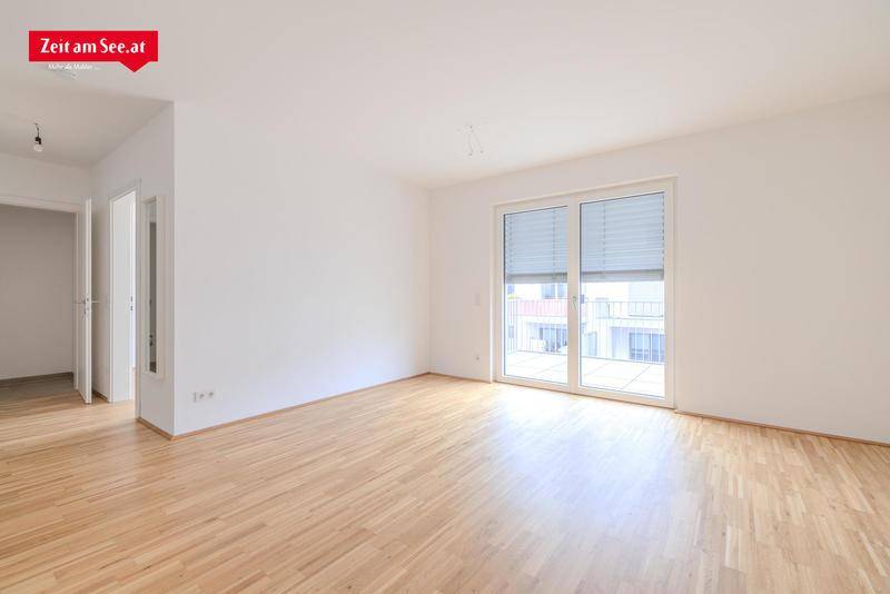 Helles, leeres Wohnzimmer mit Parkettboden und großem Fenster, das Zugang zu einem Balkon bietet.